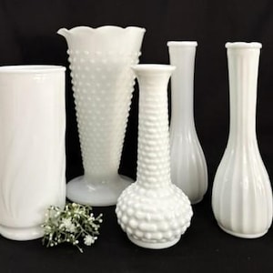 以下が含まれることがあります： さまざまな形をした5つの白いガラスの花瓶のコレクション。花瓶には、背の高い円筒形の花瓶、ホブネイルの花瓶、リブと溝のデザインの花瓶が含まれます。白い花の小さな小枝が花瓶の前にあります。