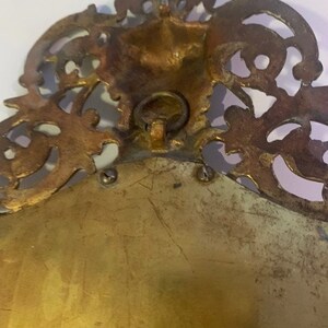 Puede incluir: Un marco de imagen de metal dorado con un dise&ntilde;o ornamentado y decorativo. El marco tiene un anillo en la parte posterior para colgar.