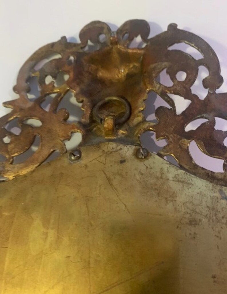 Puede incluir: Un marco de imagen de metal dorado con un dise&ntilde;o ornamentado y decorativo. El marco tiene un anillo en la parte posterior para colgar.