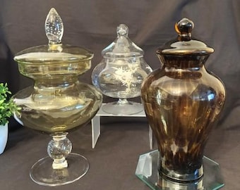 May include: Three glass apothecary jars with lids. The left jar is olive-tinted glass on a pedestal base. The centre jar is clear glass with a floral etching on a pedestal. The right jar is dark brown glass with a vertical ribbed texture.
