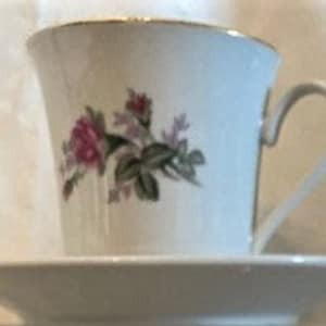 May include: A white teacup and saucer set with a floral design featuring pink roses and green leaves. The cup has a gold rim and the saucer has a plain white design.