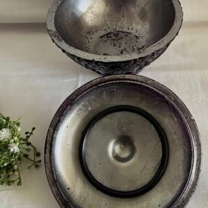 Puede incluir: Un cuenco de metal plateado y morado con un exterior texturizado y un interior liso. El cuenco tiene una tapa con una abertura circular en el centro.