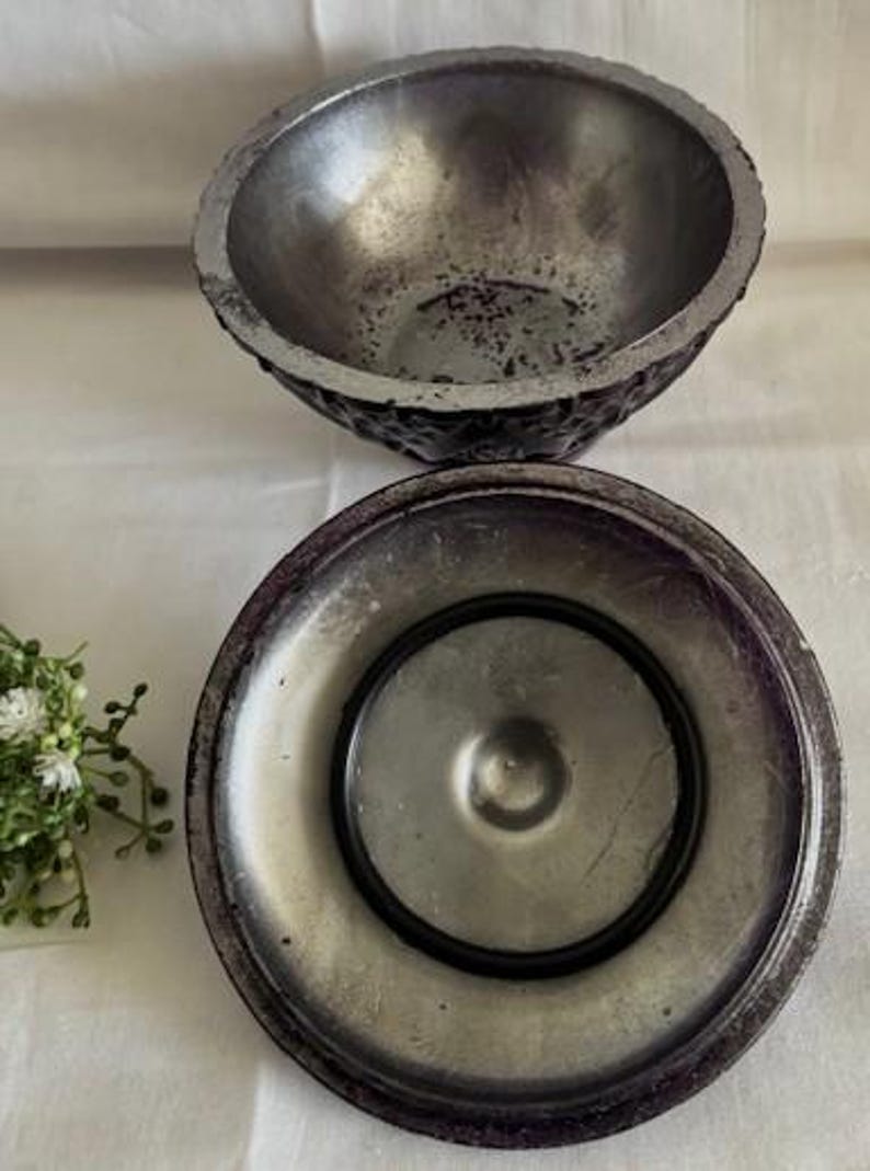 Puede incluir: Un cuenco de metal plateado y morado con un exterior texturizado y un interior liso. El cuenco tiene una tapa con una abertura circular en el centro.