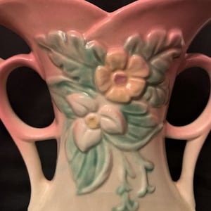 Puede incluir: Un jarrón vintage de cerámica rosa y blanco con un diseño floral. El jarrón tiene dos asas y un borde festoneado. Las flores son en tonos de rosa, amarillo y verde.