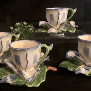 May include: Set of four blue and white teacups with green leaf-shaped saucers. Each saucer features a 3D flower design.