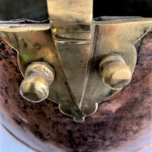 May include: Close-up of a copper pot with a brass handle and two brass knobs. The pot has a worn, aged appearance.