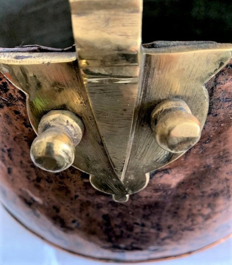 May include: Close-up of a copper pot with a brass handle and two brass knobs. The pot has a worn, aged appearance.