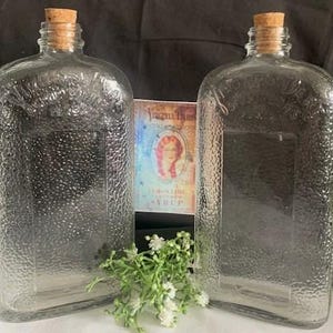 May include: Two clear glass bottles with cork stoppers. The bottles have a textured surface and are empty. The bottles are similar in size and shape.