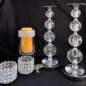 May include: An assortment of decorative candle holders and a candle. The candle holders include two tall crystal beaded candlesticks, a small crystal beaded candle holder, and a glass candle holder with a rhinestone band. The candle is orange.