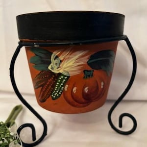 以下が含まれることがあります： 黒い縁取りと金属製のスタンドが付いた小さなテラコッタ製の鉢。鉢には、カボチャ、トウモロコシ、葉が秋の色の絵で描かれています。