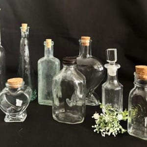 May include: A collection of clear glass bottles and jars with cork stoppers. The assortment includes various shapes and sizes, from tall, slender bottles to heart-shaped jars. Some bottles have a square or rectangular base.