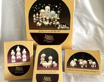 Może przedstawiać: Kolekcja miniaturowych szopek i ozdób Precious Moments. Pudełka są w kolorze beżowym z ciemnobrązowymi akcentami i zawierają ilustracje sceny Bożego Narodzenia. Tekst zawiera "Come Let Us Adore Him" i "A Trio of Porcelain Bisque Ornaments."