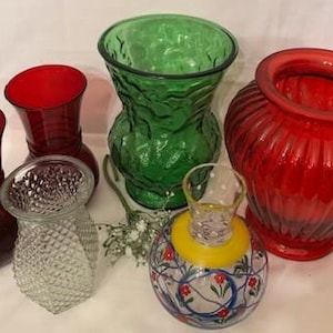 May include: A collection of colorful glass vases in various shapes and sizes. The vases include shades of red, green, and clear glass. One vase has a floral design, and another has a textured surface. The vases are arranged on a white surface.