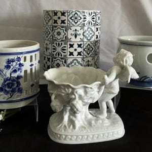 Könnte beinhalten: Eine Sammlung von Keramikobjekten, bestehend aus einer weißen Porzellanschale, die von einer Putto-Figur gehalten wird, einem zylindrischen Behälter mit blau-weißem Fliesenmuster, einem weißen Gitterhalter mit blauen Blumenmotiven und einer weißen Schale mit blauen Akzenten.