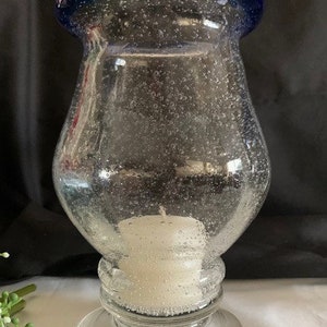 May include: A clear glass candle holder with a blue rim and a textured surface. The holder has a wide base and a narrow neck. A white candle is inside the holder.