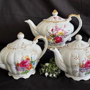 May include: Three white porcelain teapots with floral designs and gold accents. The teapots have different shapes and sizes, and each features a unique floral pattern.