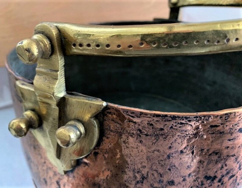May include: A copper pot with a brass handle and decorative accents. The pot has a hammered finish and is slightly dented.