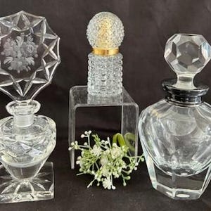 May include: Three clear glass perfume bottles with decorative stoppers. One bottle has a floral design etched into the stopper. Another has a gold band around the neck. The bottles are on a black surface.