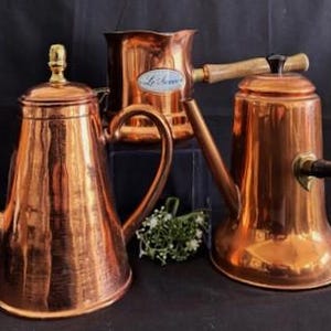May include: Three copper teapots with different styles of handles and spouts. The teapot on the left has a hammered finish, the middle teapot has a wooden handle, and the teapot on the right has a black handle.