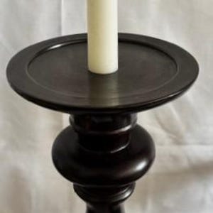 May include: A black wooden candle holder with a single white candle. The candle holder has a round base and a tall, slender stem.