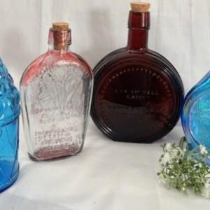 Puede incluir: Cuatro botellas de vidrio vintage con tapones de corcho. Las botellas son azules, transparentes, rojas y azul iridiscente. Las botellas tienen diferentes formas y tamaños. La botella transparente tiene una etiqueta con texto.