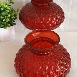 Puede incluir: Dos pantallas de lámpara de cristal rojo a juego con un patrón texturizado de puntos en relieve. Cada pantalla tiene un cuerpo redondeado y abombado con una abertura ancha y festoneada en la parte superior. El cristal es rojo intenso.