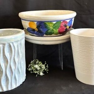 May include: Three ceramic planters of varying designs. One is a white, textured cylinder. Another is a light gray, wavy-patterned oval. The third is a bowl with a floral design of pansies in blue, yellow, and red.