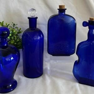 May include: Four cobalt blue glass bottles of varying shapes. One has a round stopper, one a clear crystal-like stopper, one a cork stopper, and one a violin-shaped bottle with a cork. The bottles are smooth, translucent, and deep blue.