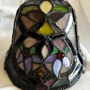 May include: A stained glass lampshade with a black border. The shade features a floral design with purple, green, white, and yellow glass pieces. The lampshade is bell-shaped and has a textured appearance.
