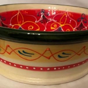 Puede incluir: Un cuenco de cerámica decorativo con un borde ancho y una base de pedestal. El interior es rojo con un diseño floral, mientras que el exterior presenta patrones amarillos, rojos y azules. El borde es verde y el cuenco tiene dos asas pequeñas.