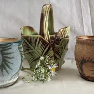 May include: Three ceramic planters in a row. The left planter is blue with a fern design. The middle planter is a green basket with a handle. The right planter is brown with a sunburst design.