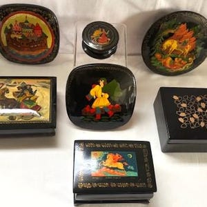Puede incluir: Una colección de siete cajas de laca negra con escenas pintadas a mano, incluyendo una troika con caballos, una figura en amarillo, un castillo, un caballo alado y motivos florales. Estos recipientes decorativos varían en forma y tamaño.