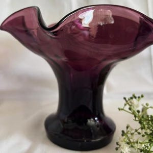 Puede incluir: Un jarr&oacute;n de vidrio morado oscuro con una abertura ancha y con volantes y una base alta y delgada.