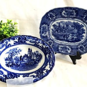 May include: Two oval ceramic platters featuring a blue and white transferware pattern. The designs depict detailed landscape scenes with buildings, trees, and water, surrounded by ornate floral borders on the scalloped edges of the dishes.