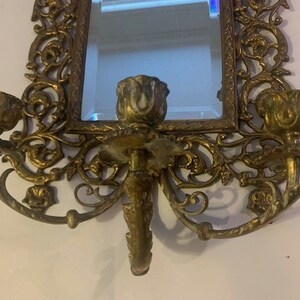 Puede incluir: Espejo de pared de lat&oacute;n adornado con candelabros a cada lado. El espejo est&aacute; rodeado por un marco decorativo con intrincados dise&ntilde;os florales. Los candelabros tienen forma de rosas.