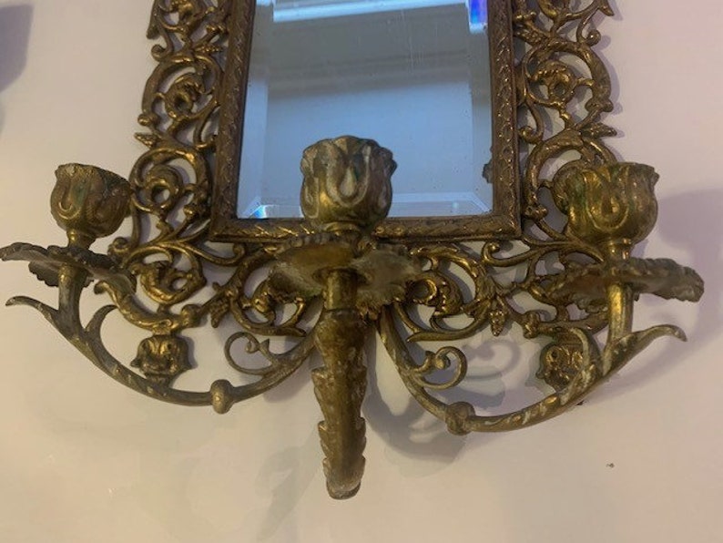 Puede incluir: Espejo de pared de lat&oacute;n adornado con candelabros a cada lado. El espejo est&aacute; rodeado por un marco decorativo con intrincados dise&ntilde;os florales. Los candelabros tienen forma de rosas.