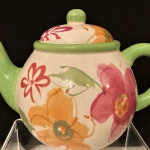 May include: A white ceramic teapot with a green lid and handle. The teapot is decorated with a floral pattern in shades of pink, orange, and green.