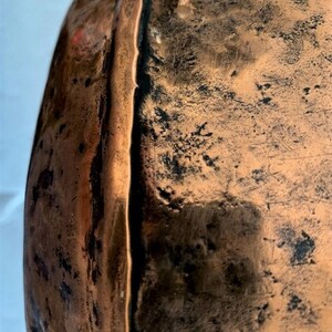 May include: Close-up of the edge of a large copper pot with a hammered finish. The copper has a dark patina and shows signs of wear.