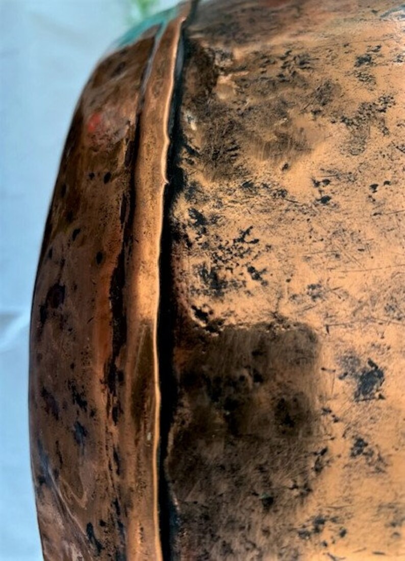 May include: Close-up of the edge of a large copper pot with a hammered finish. The copper has a dark patina and shows signs of wear.