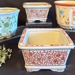 Puede incluir: Una colección de macetas de cerámica de varias formas y tamaños. Las macetas presentan diseños florales en verde, rojo y azul sobre un fondo blanco. Una maceta tiene forma hexagonal, mientras que otras son cuadradas o redondas. Adecuadas para uso interior o exterior, medidas en centímetros.