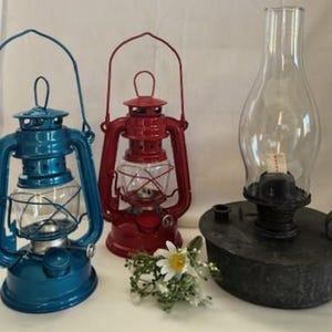 May include: Three vintage oil lanterns in a row. A teal lantern, a red lantern, and a black lantern with a glass chimney. The lanterns have glass globes and metal handles. A small bouquet of white flowers sits in front of the lanterns.