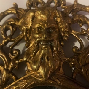 Puede incluir: Un espejo adornado en tono dorado con un rostro esculpido detallado de una criatura m&iacute;tica en el centro. El espejo est&aacute; rodeado por un marco decorativo con intrincados trabajos de volutas.