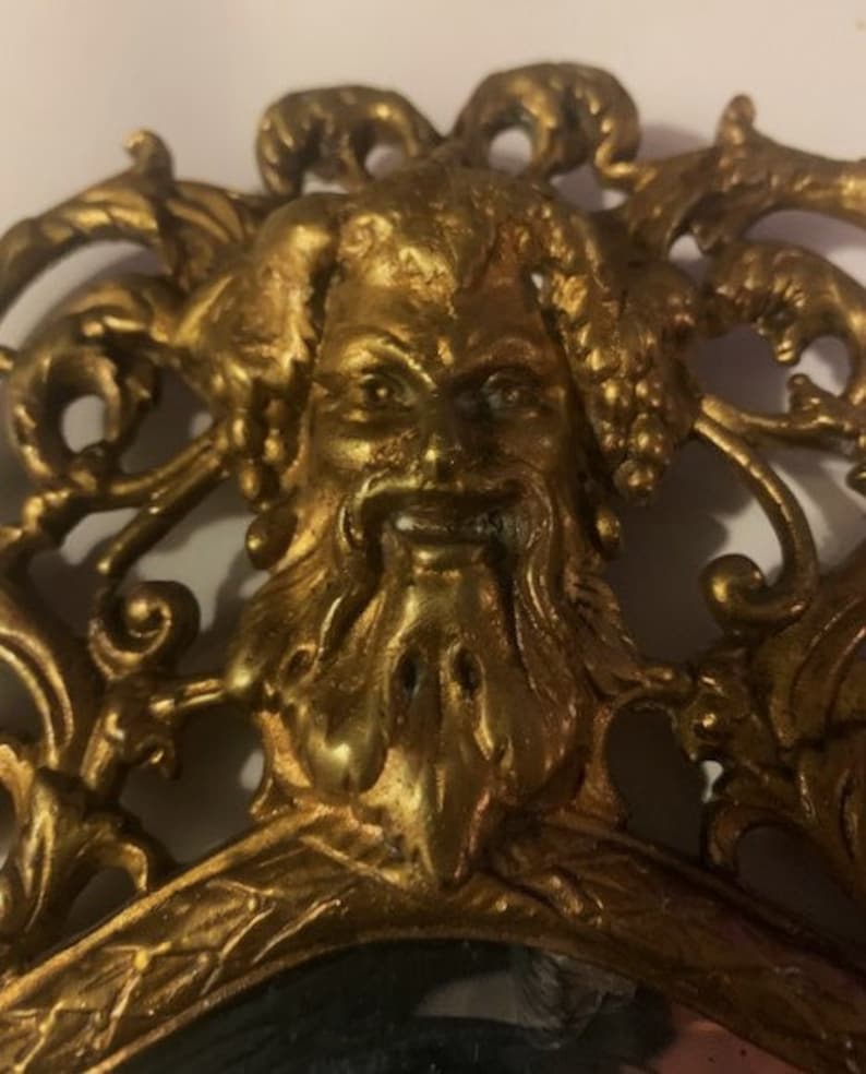 Puede incluir: Un espejo adornado en tono dorado con un rostro esculpido detallado de una criatura m&iacute;tica en el centro. El espejo est&aacute; rodeado por un marco decorativo con intrincados trabajos de volutas.
