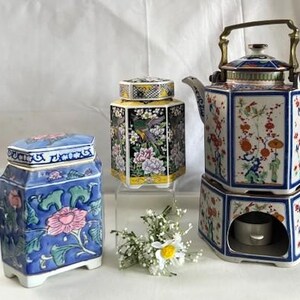 Könnte beinhalten: Eine Sammlung dekorativer Keramik-Teesets. Das Set umfasst eine blau-rosa Blumendose, eine schwarz-gelbe Blumendose und eine Teekanne mit Wärmer. Die Teekanne und der Wärmer haben ein Blumendesign mit roten, orangen und grünen Akzenten.