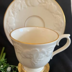May include: A white porcelain teacup and saucer with a gold rim and embossed grapevine design. The saucer has a raised rim with a gold trim.