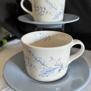 May include: Two white teacups with matching saucers. The cups and saucers are light blue. Each cup has a floral design in shades of blue and gray. The cups are on a white surface.