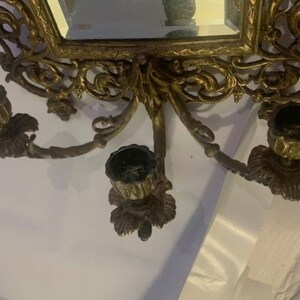 Puede incluir: Aplique de pared de metal dorado ornamentado con un espejo y tres candelabros. La aplique tiene un dise&ntilde;o floral y est&aacute; en el estilo de una ara&ntilde;a vintage.