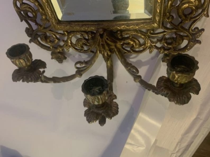 Puede incluir: Aplique de pared de metal dorado ornamentado con un espejo y tres candelabros. La aplique tiene un dise&ntilde;o floral y est&aacute; en el estilo de una ara&ntilde;a vintage.