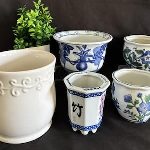 May include: A collection of five ceramic planters including one solid white pot with embossed scrollwork, one blue and white floral pot, one hexagonal pot with blue patterns and black Chinese characters, and two small pots with blue and green floral designs.