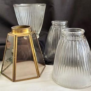 Puede incluir: Un surtido de pantallas de lámparas de vidrio vintage y una linterna decorativa. Las pantallas son transparentes con detalles acanalados verticales. La linterna tiene un marco de metal dorado y paneles de vidrio transparente. El fondo es una tela oscura.